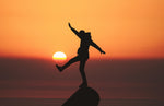 The dark profile of a person standing on one leg on a rock backlit by a glowing sunset