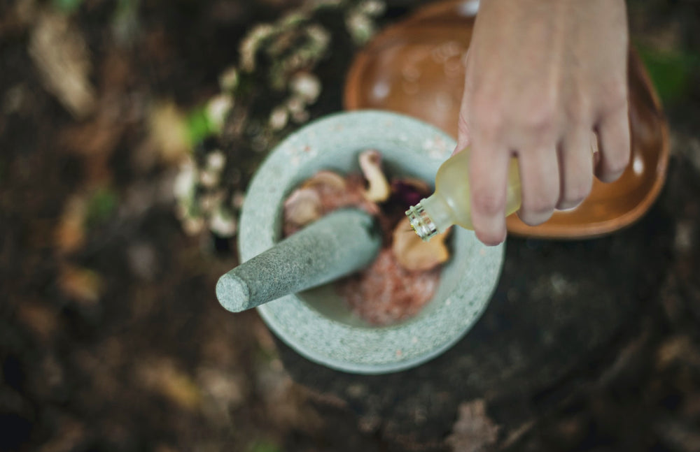 An overhead shot of a hand pouring liquid into a stone bowl with rose petals and leaves.